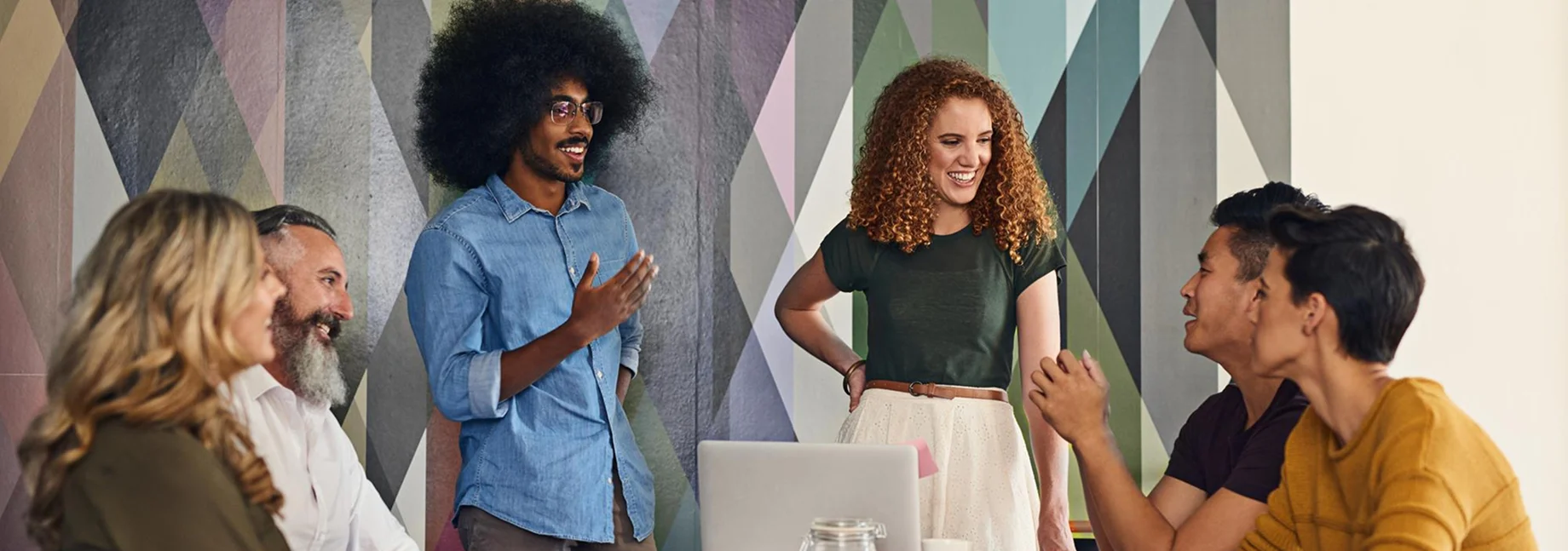 Six diverse colleagues smiling and discussing around a table with a laptop in a modern office with a geometric patterned wall.