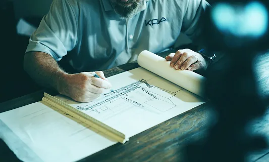 Man in a blue shirt drawing architectural plans on a wooden table with a ruler nearby.